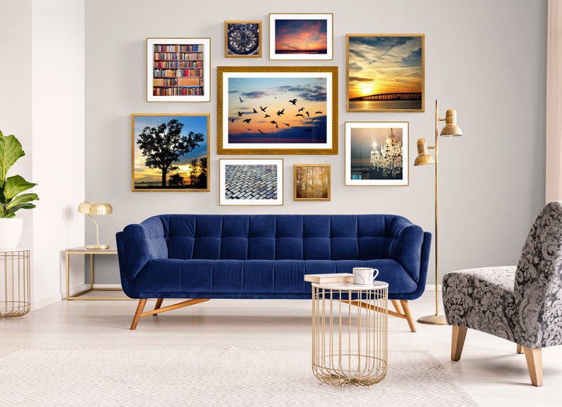 May include: A living room with a blue velvet sofa, a gold metal side table, and a gray patterned armchair. The wall is decorated with a gallery wall of framed photos, including a sunset over the ocean, a cityscape, and a chandelier.