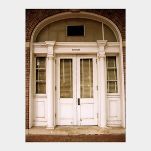May include: A white double door with a fanlight above it. The door is framed by a white archway and columns. The door has a set of blinds on each side. The door is set in a brick wall.