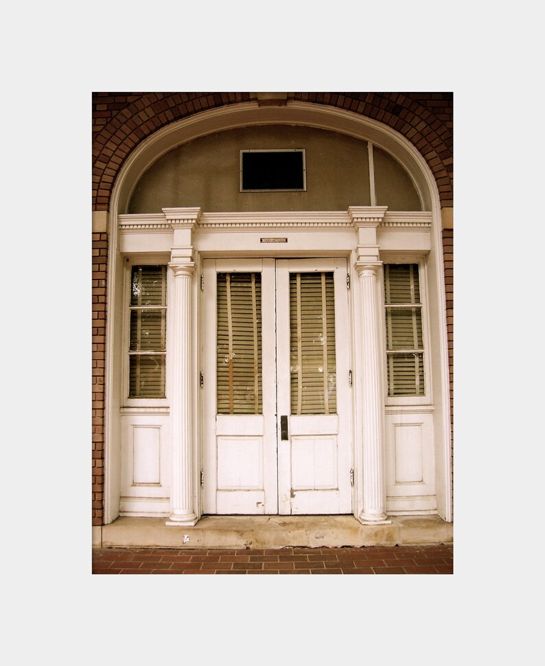 May include: A white double door with a fanlight above it. The door is framed by a white archway and columns. The door has a set of blinds on each side. The door is set in a brick wall.