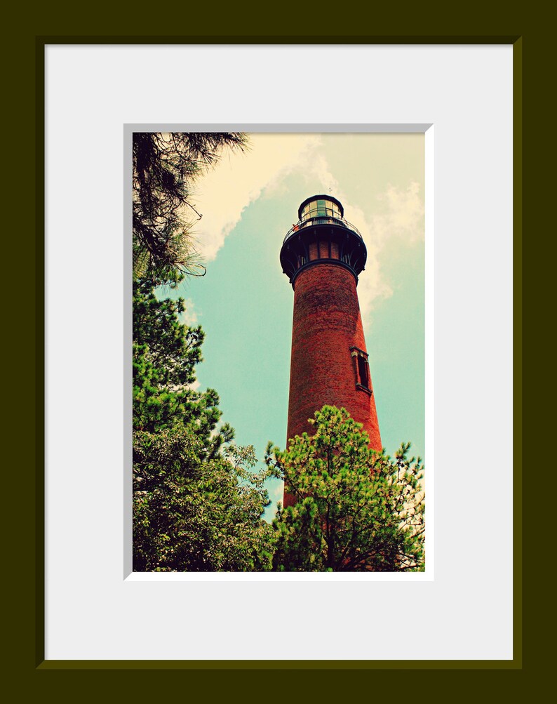May include: A red brick lighthouse with a black roof and a white window stands tall against a blue sky with white clouds. Green trees surround the base of the lighthouse.