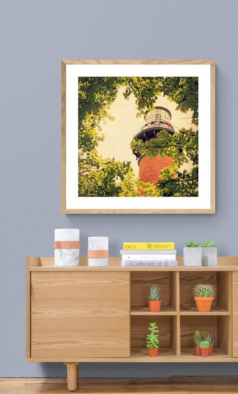 May include: A framed photograph of a red brick lighthouse with a white roof, viewed from below, surrounded by green trees. The lighthouse is tall and slender, with a white railing around the top. The trees are in full leaf, and the sky is a pale blue.