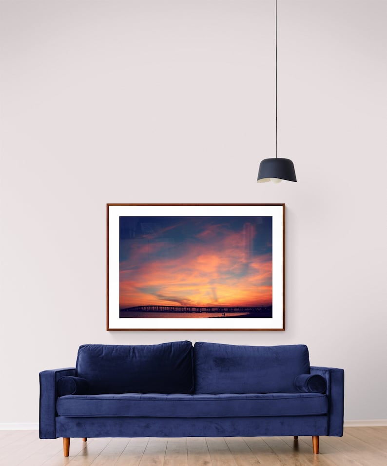 May include: A framed photograph of a sunset over a beach with a bridge in the distance. The sky is a vibrant orange and pink, and the water is a deep blue. The photo is hanging on a white wall above a blue velvet sofa.