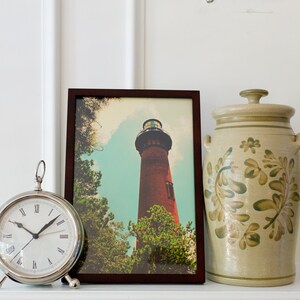 May include: A framed photograph of a red brick lighthouse against a blue sky with white clouds. The lighthouse is surrounded by green trees. The photograph is sitting on a white shelf with a silver clock and a beige ceramic jar with green floral designs.