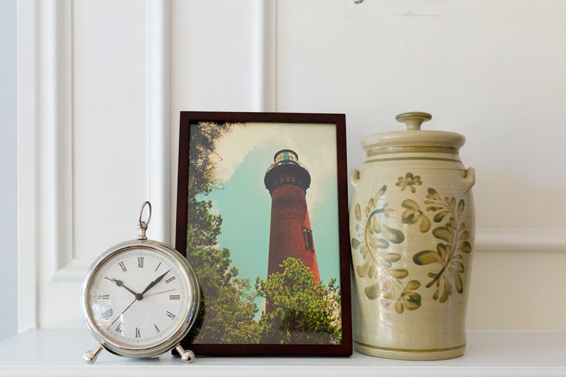 May include: A framed photograph of a red brick lighthouse against a blue sky with white clouds. The lighthouse is surrounded by green trees. The photograph is sitting on a white shelf with a silver clock and a beige ceramic jar with green floral designs.