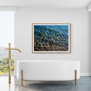 Pu&ograve; includere: Una vasca da bagno autoportante bianca con gambe in metallo dorato in un bagno moderno con una grande finestra che si affaccia su un paesaggio tropicale. Una fotografia incorniciata di acqua increspata &egrave; appesa al muro sopra la vasca da bagno.