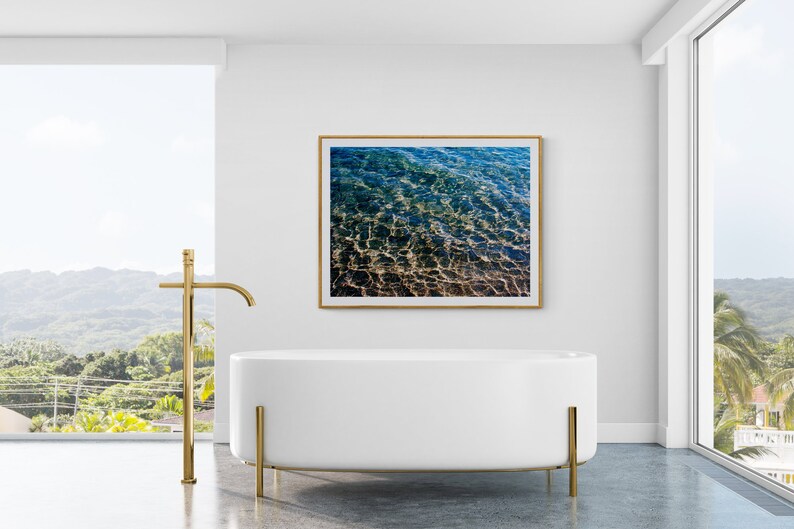 Pu&ograve; includere: Una vasca da bagno autoportante bianca con gambe in metallo dorato in un bagno moderno con una grande finestra che si affaccia su un paesaggio tropicale. Una fotografia incorniciata di acqua increspata &egrave; appesa al muro sopra la vasca da bagno.