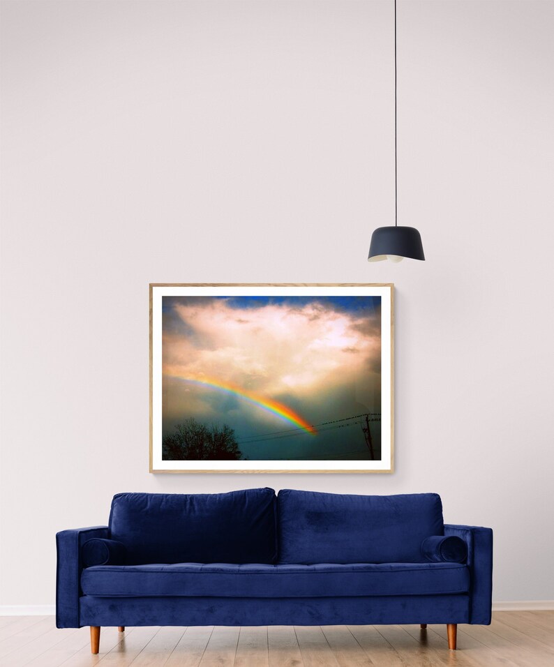 May include: A framed photograph of a rainbow in the sky with a single cloud in the centre of the frame. The rainbow is a vibrant arc of colour against a blue sky. The photograph is hanging on a white wall above a blue velvet sofa.