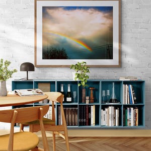 May include: A framed photograph of a rainbow in the sky with a large, fluffy cloud. The photo is hanging on a white brick wall above a blue bookshelf filled with books and decorative items.