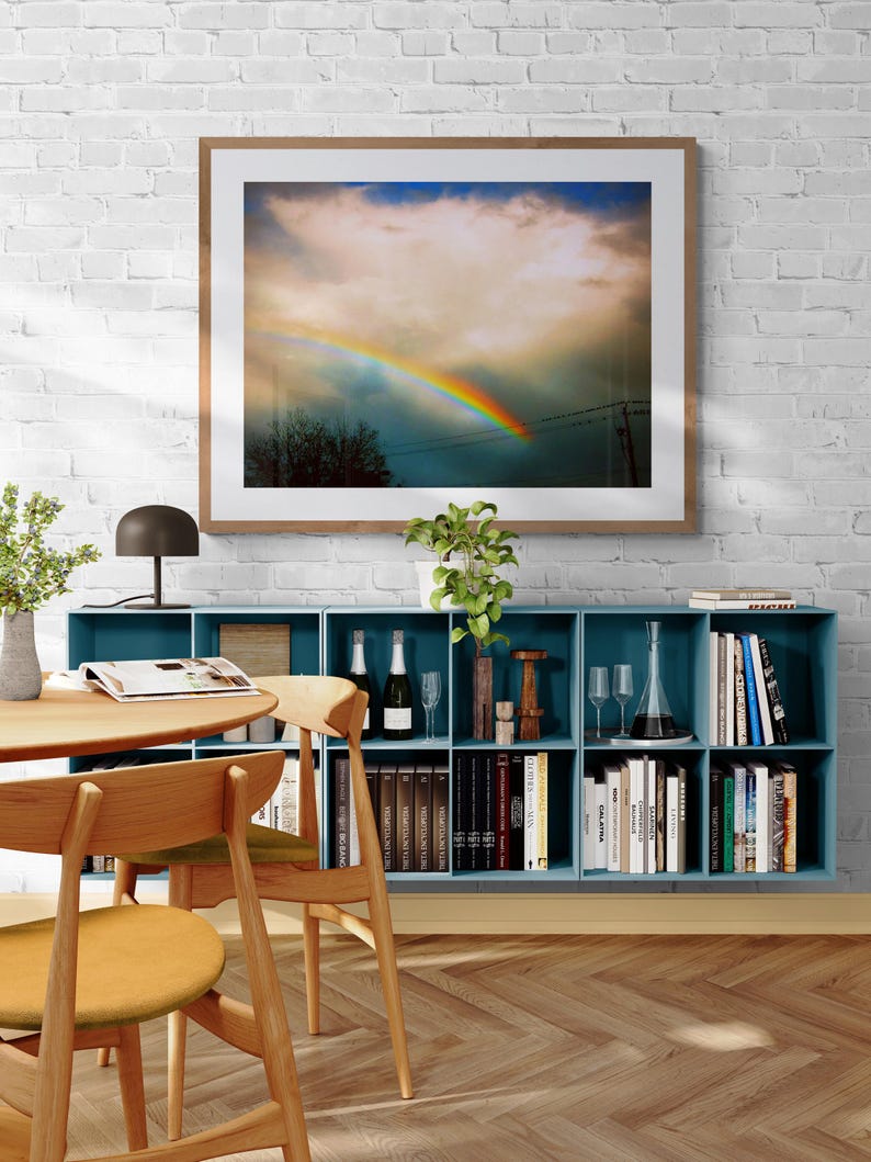 May include: A framed photograph of a rainbow in the sky with a large, fluffy cloud. The photo is hanging on a white brick wall above a blue bookshelf filled with books and decorative items.