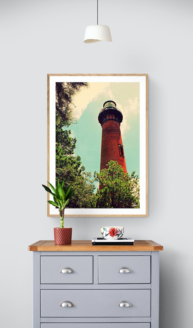 May include: A framed photograph of a red brick lighthouse with a black roof and a white trim. The lighthouse is surrounded by green trees and a blue sky with white clouds. The photograph is hanging on a white wall above a gray dresser with a wooden top.
