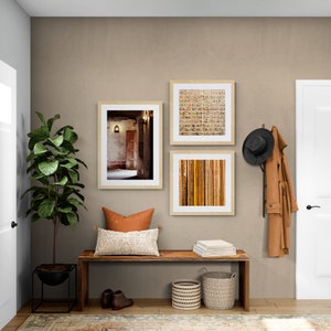 May include: A wooden bench with a brown cushion and a white throw pillow sits in a hallway with three framed pictures on the wall. The pictures are of a brick building, a striped pattern, and a brick wall with a door. There is a hat rack with a brown hat and a brown coat hanging on it. There are two woven baskets on the floor next to the bench.