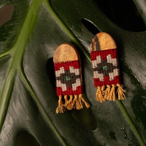 May include: Two handmade earrings featuring a wooden top and a woven pattern in red, white, black, and green. The earrings have a rectangular shape with a fringe of yellow threads. They are presented on a large green leaf, creating a natural backdrop.