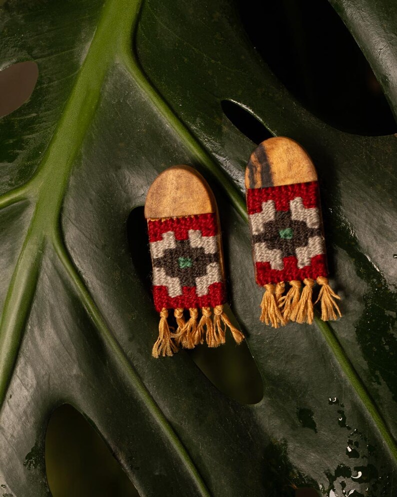 May include: Two handmade earrings featuring a wooden top and a woven pattern in red, white, black, and green. The earrings have a rectangular shape with a fringe of yellow threads. They are presented on a large green leaf, creating a natural backdrop.