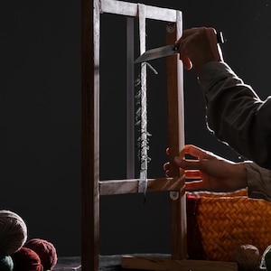 May include: A wooden weaving loom against a dark backdrop. A person is using a knife to cut a strand of yarn. Several balls of yarn in various colours are in the foreground, along with a woven basket.