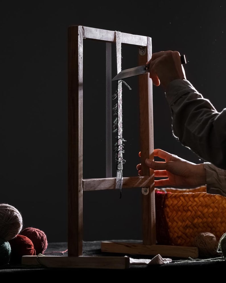 May include: A wooden weaving loom against a dark backdrop. A person is using a knife to cut a strand of yarn. Several balls of yarn in various colours are in the foreground, along with a woven basket.