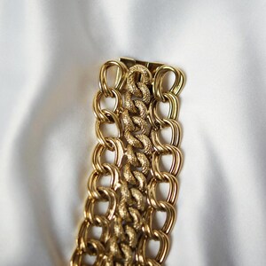 May include: A gold-toned chain bracelet with a textured center section and large, oval-shaped links. The bracelet is displayed on a white, silky background, highlighting its shiny, metallic finish. The design is bold and eye-catching.