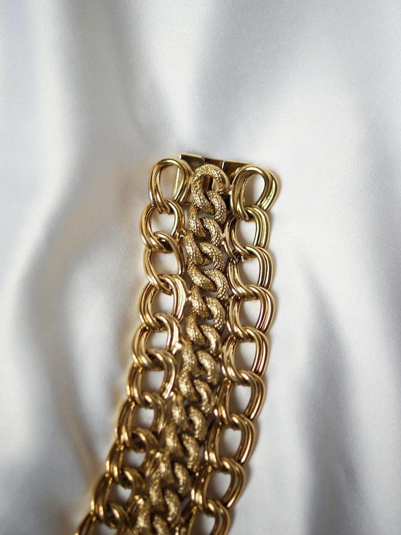 May include: A gold-toned chain bracelet with a textured center section and large, oval-shaped links. The bracelet is displayed on a white, silky background, highlighting its shiny, metallic finish. The design is bold and eye-catching.