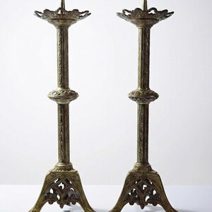May include: Two antique brass candle holders. Each has a decorative base, a long, detailed stem, and a floral-shaped top with a central spike. The candle holders are displayed against a white background.