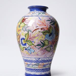 Puede incluir: Un jarrón de cerámica con cuerpo bulboso y cuello estrecho, que presenta un patrón floral pintado a mano en tonos azul, amarillo, verde y rosa sobre un fondo violeta claro, con bandas geométricas de rayas cruzadas azules cerca de la base.