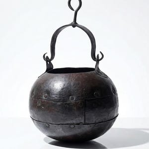 May include: A dark brown, spherical metal cauldron with a curved handle and a hook for hanging. The cauldron is constructed from riveted metal plates, giving it a rustic appearance. The handle is attached to the cauldron with curved brackets.