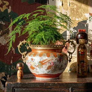 Peut inclure: Un grand pot en céramique orné de motifs floraux et d'oiseaux, rempli d'une plante verte. Deux petites poupées en bois et une table en bois sombre complètent la scène, le tout sur un fond à motifs.