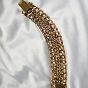 May include: A gold-toned chain bracelet with multiple rows of linked chains. The bracelet is secured with a rectangular clasp. The chains are arranged on a white, textured surface.
