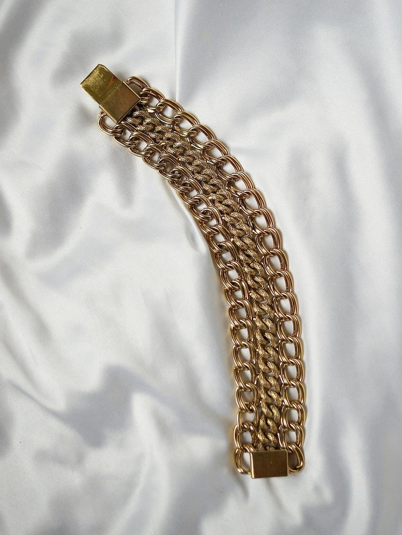 May include: A gold-toned chain bracelet with multiple rows of linked chains. The bracelet is secured with a rectangular clasp. The chains are arranged on a white, textured surface.