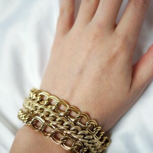 May include: Gold-toned chain bracelets in varying designs are stacked on a wrist. The bracelets have different link styles, including oval and textured patterns. The jewelry is displayed against a white background, highlighting the gold color.