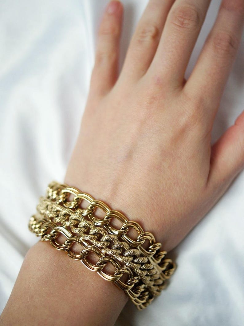 May include: Gold-toned chain bracelets in varying designs are stacked on a wrist. The bracelets have different link styles, including oval and textured patterns. The jewelry is displayed against a white background, highlighting the gold color.