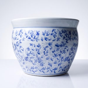 May include: A round ceramic planter featuring a white base with a hand-painted blue floral vine pattern. The vessel has a wide, open top with a smooth, curved rim and a tapered base, showcasing a classic blue and white colour scheme throughout.