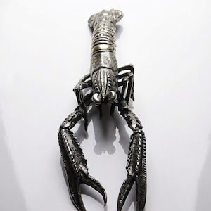 Può includere: Una dettagliata statuetta di aragosta in metallo argentato. Il crostaceo è raffigurato con dettagli intricati sul corpo, sulle chele e sulle zampe. Le chele dell'aragosta sono aperte, su sfondo bianco.
