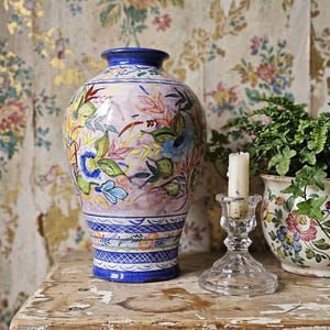 Könnte beinhalten: Eine Keramikvase mit handgemaltem Blumenmuster in Blau-, Rosa-, Gelb- und Grüntönen. Die Vase hat einen bauchigen Körper, einen schmalen Hals und einen blauen Rand. Sie steht neben einem Kerzenhalter aus Klarglas mit einer weißen Kerze.