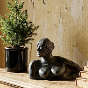 Może przedstawiać: Brązowa rzeźba leżącej postaci o spokojnym wyrazie twarzy. Rzeźba stoi na drewnianej powierzchni, obok małego drzewka iglastego w czarnej doniczce. Tło przedstawia teksturowaną ścianę.