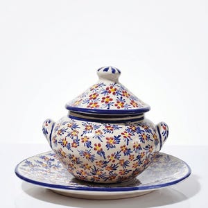 Op de afbeelding: Een keramische soepterrine met bijpassend deksel en schotel. De terrine en het deksel hebben een bloemmotief in rood, blauw en geel op een crèmekleurige achtergrond. De rand is blauw.