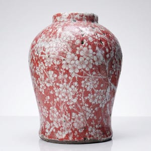 Peut inclure: Un vase en céramique au corps arrondi et au col court, présentant une glaçure rouge vif ornée d'un motif floral blanc de fleurs de cerisier. La surface a une finition texturée et légèrement irrégulière avec de petites taches sombres.