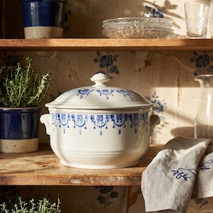 Op de afbeelding: Een witte keramische ovenschaal met een blauw bloemmotief en een bijpassende deksel staat op een houten plank. De schaal heeft twee handvatten en is omgeven door andere keukenspullen, waaronder glazen en een linnen servet.