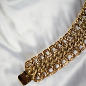 May include: A gold-toned chain bracelet with a textured center chain and a rectangular clasp. The bracelet features three rows of interlocking links, creating a bold and stylish accessory. The background is a white, silky fabric.
