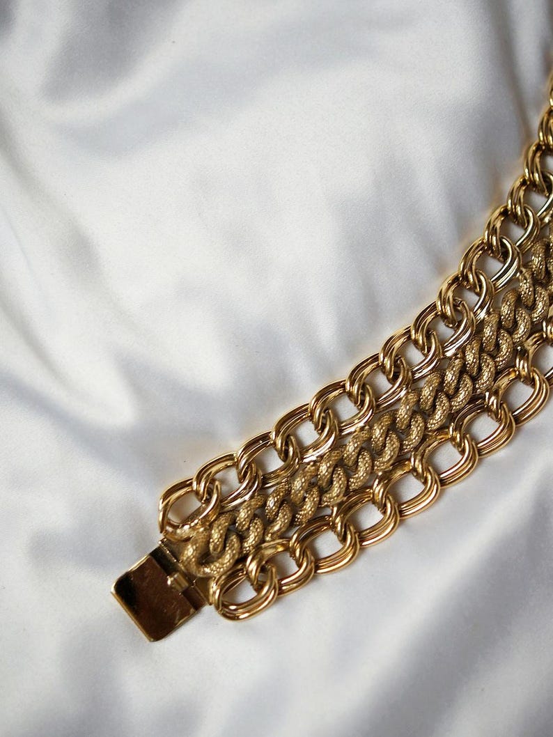 May include: A gold-toned chain bracelet with a textured center chain and a rectangular clasp. The bracelet features three rows of interlocking links, creating a bold and stylish accessory. The background is a white, silky fabric.