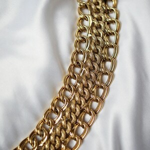 May include: A gold-toned chain necklace with a textured, interwoven design. The necklace is composed of multiple strands of linked chains, creating a bold and eye-catching accessory. The background is a white, silky fabric.