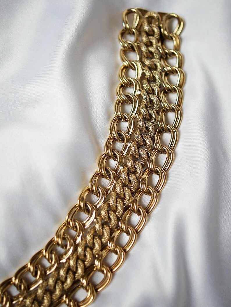 May include: A gold-toned chain necklace with a textured, interwoven design. The necklace is composed of multiple strands of linked chains, creating a bold and eye-catching accessory. The background is a white, silky fabric.