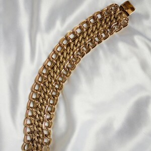 May include: A gold-toned bracelet with a chain link design. The bracelet features multiple rows of interlocking links, creating a bold and textured appearance. The clasp is rectangular and matches the gold color of the bracelet.