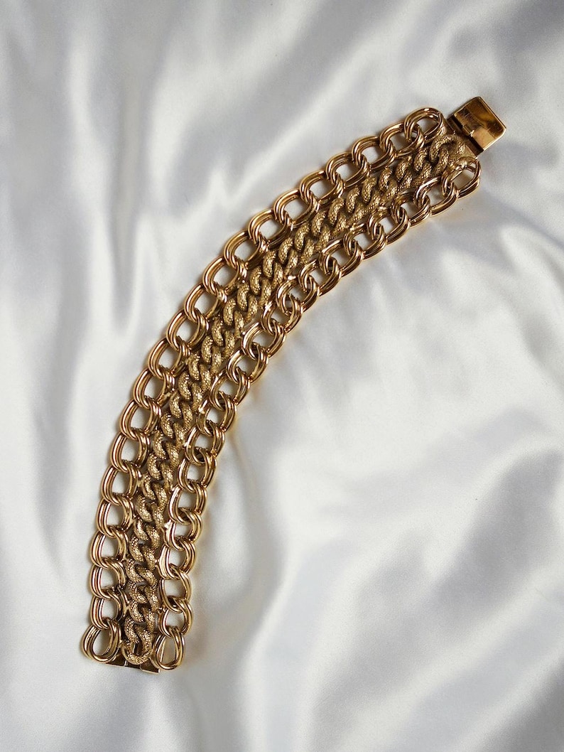 May include: A gold-toned bracelet with a chain link design. The bracelet features multiple rows of interlocking links, creating a bold and textured appearance. The clasp is rectangular and matches the gold color of the bracelet.