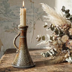 Op de afbeelding: Een brandende kaars in een getextureerde, bruine keramische kandelaar met een handvat. De kaars is crèmekleurig en de houder heeft een gespikkelde, rustieke uitstraling. Een boeket droogbloemen en pampasgras staat in de buurt.