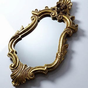 Puede incluir: Espejo dorado y ornamentado con un marco decorativo y con volutas. El espejo tiene una forma curva e irregular y está sobre un fondo blanco. El marco presenta tallas detalladas y un acabado texturizado, de estética antigua.