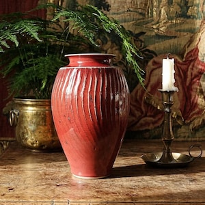 Può includere: Un vaso alto in ceramica rossa con linee verticali strutturate. Il vaso è su un tavolo di legno, con un candelabro in ottone e una pianta in vaso sullo sfondo. Il vaso ha una base larga e un collo stretto.