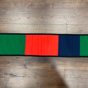 May include: A long, rectangular fabric table runner featuring a color-blocked design. The runner is divided into four vertical sections of green, bright orange, dark blue, and green, all bordered by a thin black trim along the edges.