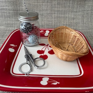 May include: A red and white ceramic tray with a cherry design. The tray has a glass jar with colorful yarn balls, a small wicker basket, and a pair of silver scissors.