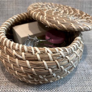 May include: A round, woven basket with a lid, made of natural-colored straw and white accents. The lid is partially open, revealing a small box and glass jars with pink lids inside. The basket sits on a gray surface.