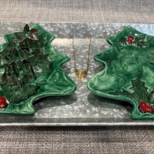 May include: Two green ceramic dishes shaped like Christmas trees with red holly berries, a set of silver metal cookie cutters nested on the left dish, and a small gold-toned metal serving tong positioned between the two dishes on a tray.