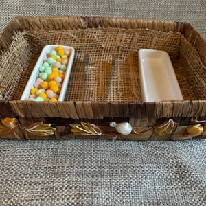 May include: A woven basket with two white rectangular dishes. One dish contains green and orange candy. The basket has a brown and gold design.